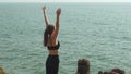 young woman stands in Urdhva Hastasana on a rock with the endless ocean below