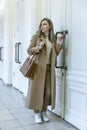 A young woman stands at a white door. Beautiful smiling blonde in a stylish beige coat. Full height. Purchase, rental of real Royalty Free Stock Photo