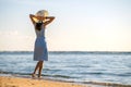 Young woman in straw hat and a dress standing alone on empty sand beach at sea shore. Lonely tourist girl looking at horizon over Royalty Free Stock Photo