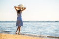 Young woman in straw hat and a dress standing alone on empty sand beach at sea shore. Lonely tourist girl looking at horizon over Royalty Free Stock Photo