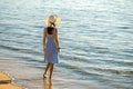 Young woman in straw hat and a dress standing alone on empty sand beach at sea shore. Lonely tourist girl looking at horizon over Royalty Free Stock Photo