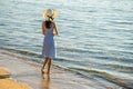 Young woman in straw hat and a dress standing alone on empty sand beach at sea shore. Lonely tourist girl looking at horizon over Royalty Free Stock Photo