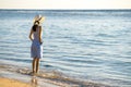 Young woman in straw hat and a dress standing alone on empty sand beach at sea shore. Lonely tourist girl looking at horizon over Royalty Free Stock Photo