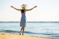 Young woman in straw hat and a dress standing raising hands on empty sand beach at sea shore. Lonely tourist girl looking at Royalty Free Stock Photo