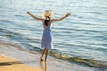 Young woman in straw hat and a dress standing raising hands on empty sand beach at sea shore. Lonely tourist girl looking at Royalty Free Stock Photo