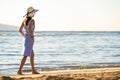 Young woman in straw hat and a dress walking alone on empty sand beach at sea shore. Lonely tourist girl looking at horizon over Royalty Free Stock Photo