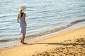 Young woman in straw hat and a dress walking alone on empty sand beach at sea shore. Lonely tourist girl looking at horizon over Royalty Free Stock Photo