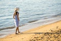 Young woman in straw hat and a dress walking alone on empty sand beach at sea shore. Lonely tourist girl looking at horizon over Royalty Free Stock Photo