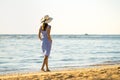 Young woman in straw hat and a dress walking alone on empty sand beach at sea shore. Lonely tourist girl looking at horizon over Royalty Free Stock Photo