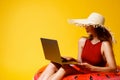 Young woman in straw sits in inflatable ring works on laptop on yellow background. freelance. Travel