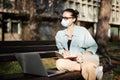 A young woman student in a facial protective mask sitting on a bench in the park and typing on her laptop on a sunny day Royalty Free Stock Photo