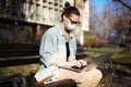 A young woman student in a facial protective mask sitting on a bench in the park and typing on her laptop on a sunny day Royalty Free Stock Photo