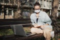 A young woman student in a facial protective mask sitting on a bench in the park and typing on her laptop on a sunny day Royalty Free Stock Photo