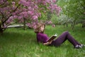 Young woman, student, reads a book with interest, lying under a tree on a spring day in park Royalty Free Stock Photo