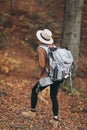 Young woman in a stylish hat and travel bag on her shoulders, looking around at the charming autumn forest Royalty Free Stock Photo