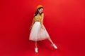 Young woman in stylish orange hat and bright blouse is dancing on red background. Girl in white skirt and sneakers Royalty Free Stock Photo