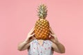 Young woman in summer clothes hold in hands hiding covering face with fresh ripe pineapple fruit on pink pastel