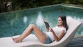 Young woman in summer clothes lies on deck chair and reads book near pool