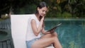 Young woman in summer clothes sits on sunbed, reads book near pool and yawns