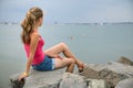 Young woman in summer clothes sitting on big boulders on sea shore looking on horizon