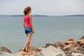 Young woman in summer clothes standing on big boulders on sea shore looking on horizon