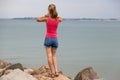 Young woman in summer clothes standing on big boulders on sea shore looking on horizon