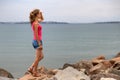 Young woman in summer clothes standing on big boulders on sea shore looking on horizon