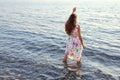 Young woman in summer dress stands barefoot in water and raising her hand up looks towards sunset Royalty Free Stock Photo