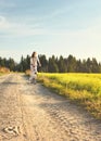 Young woman in summer light clothes rides bicycle on dusty road towards camera, afternoon sun shines to fields and forest in Royalty Free Stock Photo