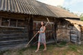 A young woman in a summer retro sarafan dress goes around the yard along the sheds and old rural buildings with rakes on the shoul