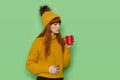 Young woman in sweater and hat with pompom is holding cup and blowing