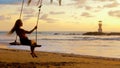 Young woman swinging sitting on swing at palm tree on sea beach in sunset lights, lighthouse on background. Happy girl Royalty Free Stock Photo