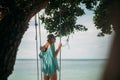 A young woman is swinging on a swing in the shade of a big tree by the sea. Royalty Free Stock Photo