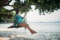 A young woman is swinging on a swing in the shade of a big tree by the sea. Royalty Free Stock Photo