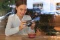 Young woman taking photo of cup with coffee at cafe, view through window. Creative hobby Royalty Free Stock Photo