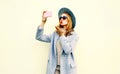 Young woman taking selfie picture by smartphone blowing red lips sending sweet air kiss in pink coat jacket, round hat Royalty Free Stock Photo