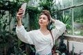 Young woman taking selfie walking in tropical greenhouse using smartphone. Modern girl wearing summer outfit