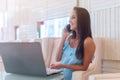 Young woman talking on mobile phone and smiling, sitting in front of the laptop in white hotel lobby Royalty Free Stock Photo