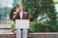 Young woman talking on the phone and typing on a laptop on street.