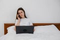 Young woman talking on phone using laptop sit on comfortable bed at home, female customer make mobile call confirming online Royalty Free Stock Photo