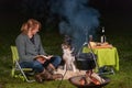 Young woman talks to her Australian Shepherd outside by a campfire. At dusk. Bread, cheese and wine on the table Royalty Free Stock Photo