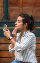 Young woman wearing casual clothes putting on makeup sitting on a park bench