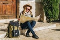 Young woman tourist sits on street on steps, looking for path on map, next to her backpack. Girl uses digital gadget.