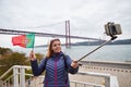 Young woman tourist take selfies on the background of famous iron 25th of April bridge and holding the flag of Portugal in hands Royalty Free Stock Photo