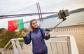 Young woman tourist take selfies on the background of famous iron 25th of April bridge and holding the flag of Portugal in hands Royalty Free Stock Photo