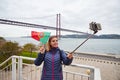 Young woman tourist take selfies on the background of famous iron 25th of April bridge and holding the flag of Portugal in hands Royalty Free Stock Photo