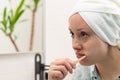 A young woman with a towel on her head in a bright bathroom with a toothbrush in her hand brushes her teeth.