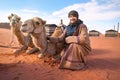 Young woman in traditional Bedouin coat - bisht - and headscarf crouching next to two camels laying on red desert ground, smiling Royalty Free Stock Photo