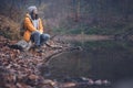 Young woman traveler in a yellow jacket and hat sitting on a log by the pond in the autumn misty forest Royalty Free Stock Photo