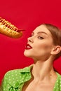 Young woman with trendy makeup eating spicy hotdog with mustard and chilli over vivid red background. Pepper instead of